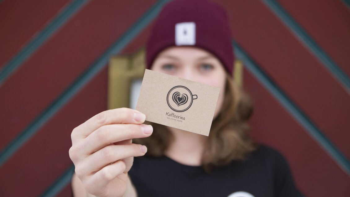 Eine Person hält eine Kaffeekarte mit einem Herz und einer Tasse als Logo. Der Hintergrund ist farbenfroh und die Person trägt eine Mütze. | © Katharina Zeiler
