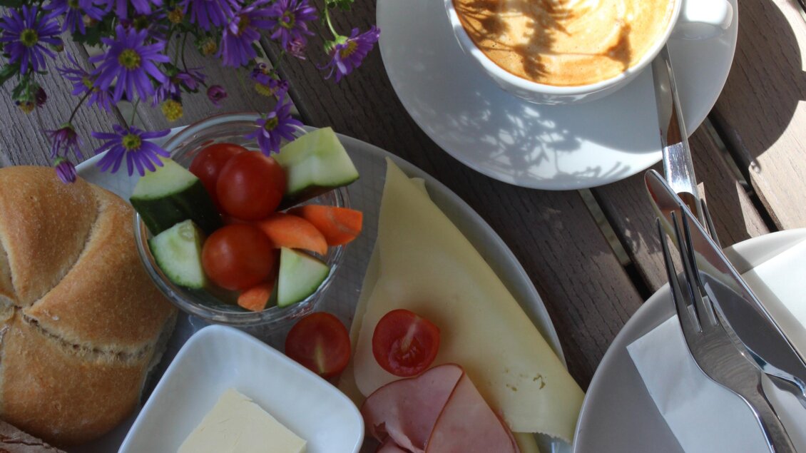 Ein schönes Frühstück mit frischen Brötchen, Käse, Schinken, Gemüse und einer Tasse Kaffee. Im Hintergrund blühen lila Blumen. | © Katharina Zeiler
