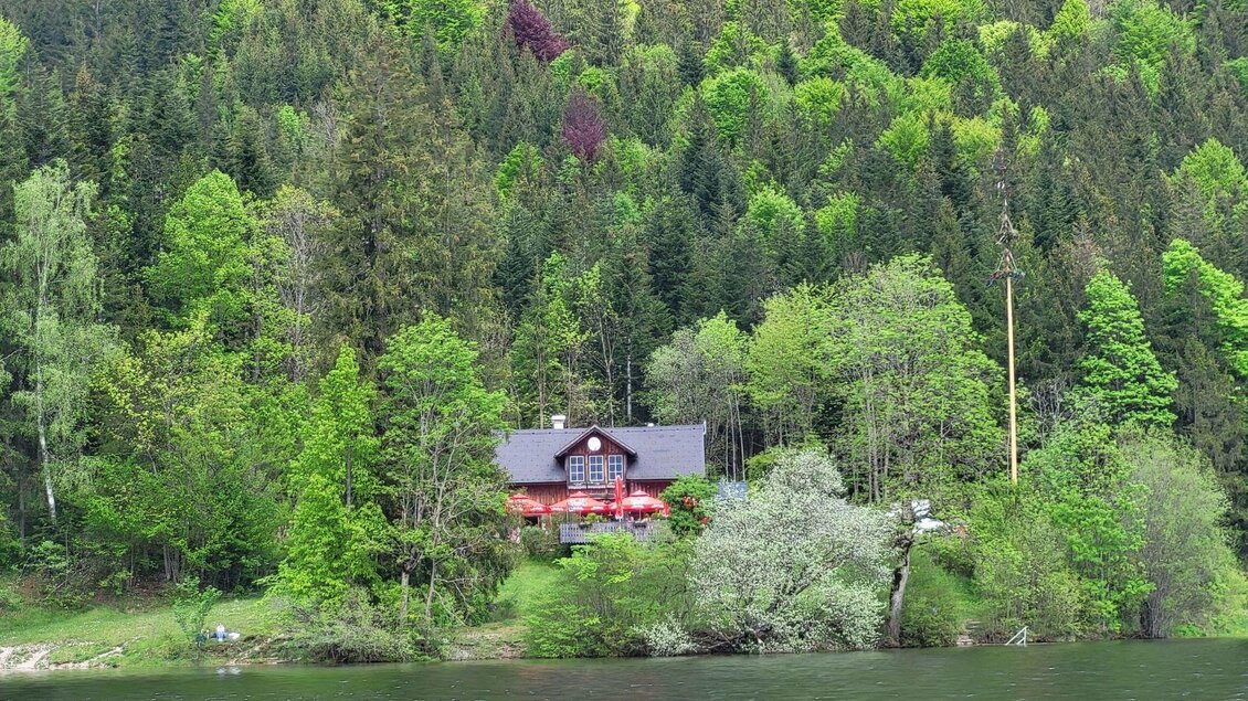 Ein charmantes Haus am Ufer eines Sees, umgeben von üppigem Grün und hohen Bäumen. Die Landschaft strahlt Ruhe und Naturverbundenheit aus. | © Petra Kirchschlager