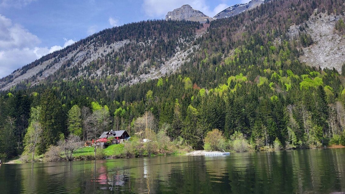 Ein ruhiger See umgeben von grünen Wäldern und Bergen. Im Vordergrund ist ein kleines, rotes Haus zu sehen. | © Petra Kirchschlager