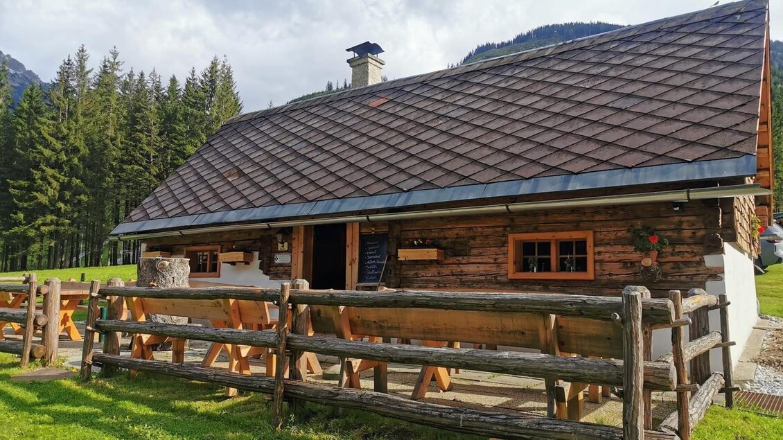 Eine gemütliche Holzhütte umgeben von grünen Wiesen und Tannenbäumen. Die Hütte hat ein steiles Dach und große Fenster. | © Kainrath
