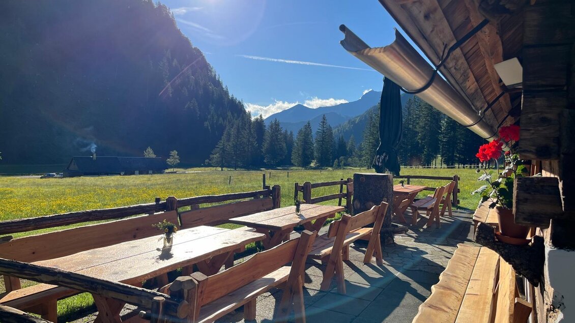 Eine idyllische Almhütte mit Holzmöbeln und einem Blick auf grüne Wiesen und Berge. Die Sonne scheint strahlend über der Landschaft. | © Kainrath