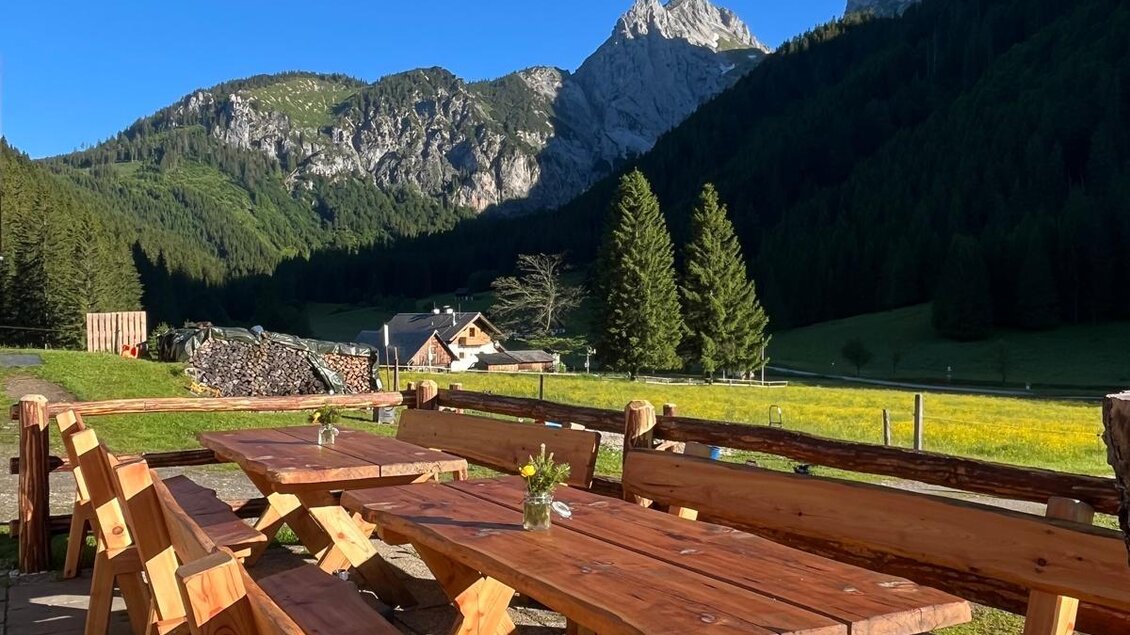 Eine einladende Holzterrasse mit langen Bänken und Tischen inmitten der Berge. Der klare blaue Himmel und die grünen Wiesen schaffen eine ruhige Atmosphäre. | © Kainrath