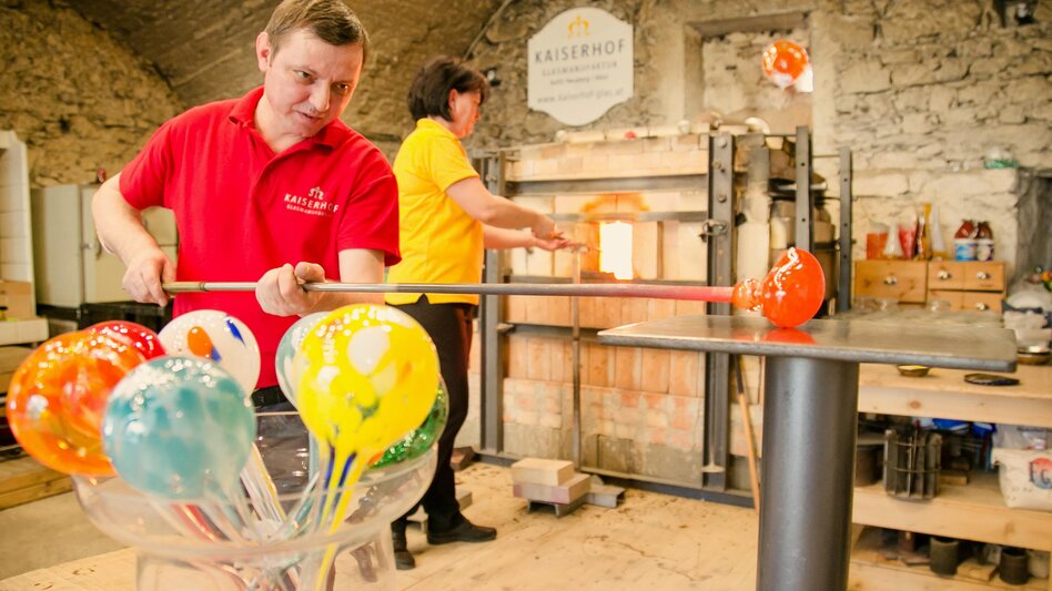 Ein Glasbläser arbeitet an einem bunten Glasobjekt in einer Werkstatt. Im Hintergrund ist eine weitere Person mit einem Ofen sichtbar. | © Kaiserhof Glasmanufaktur