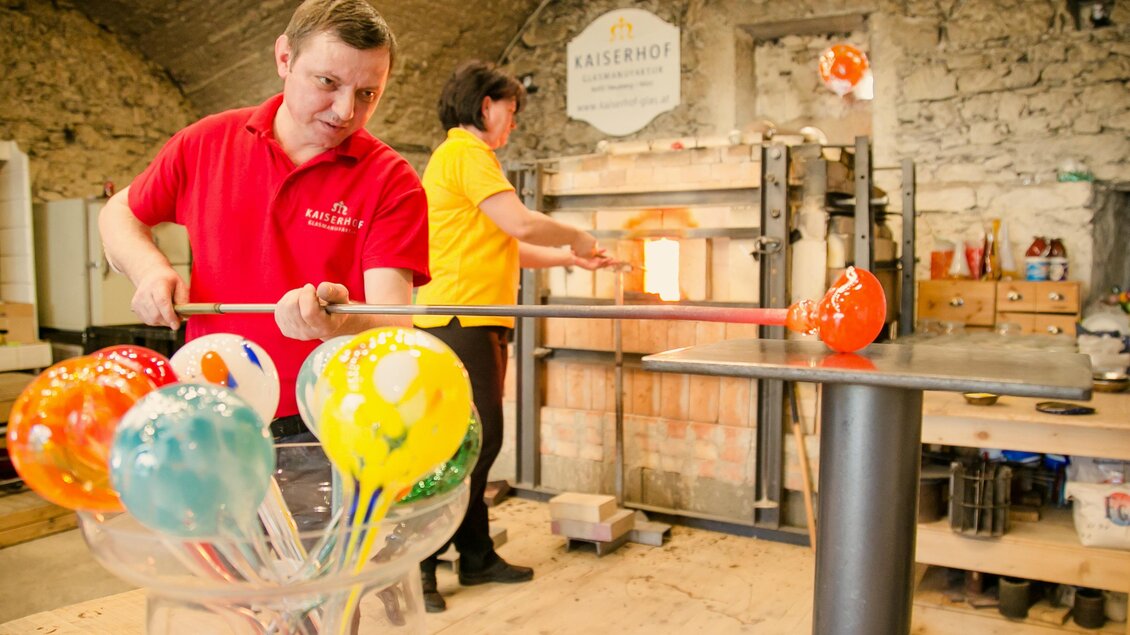 Ein Glasbläser arbeitet an einem bunten Glasobjekt in einer Werkstatt. Im Hintergrund ist eine weitere Person mit einem Ofen sichtbar. | © Kaiserhof Glasmanufaktur