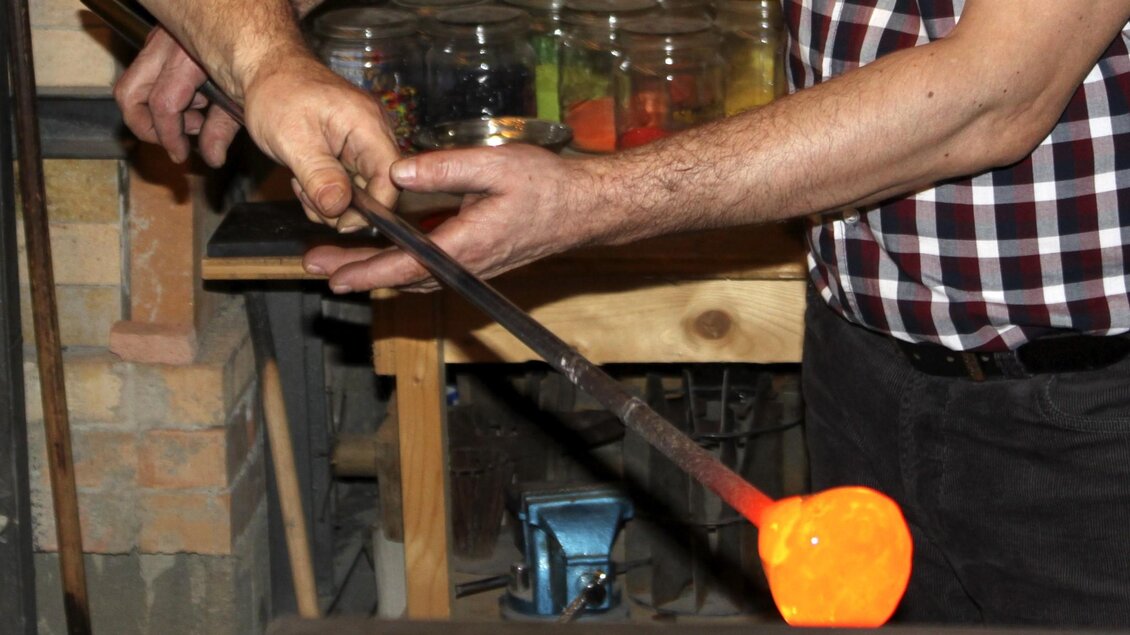 Ein Handwerker formt glühendes Glas mit einem Werkzeug. Im Hintergrund sind verschiedene Farben und Materialien sichtbar. | © Kaiserhof Glasmanufaktur