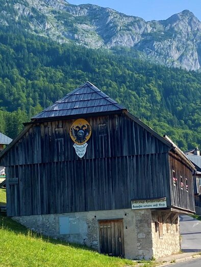 Ein traditionelles Holzhaus auf einer Wiese mit Bergen im Hintergrund. Es ist ein klarer, sonniger Tag. | © Bettina Scheck
