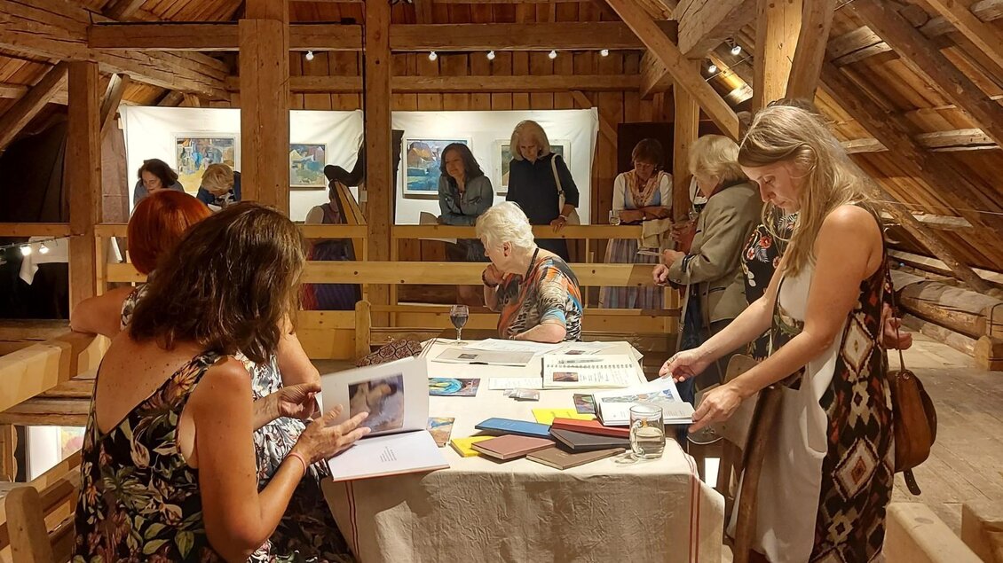 Eine Gruppe von Frauen betrachtet Kunstwerke in einem gemütlichen Raum mit Holzdecke. Einige Frauen sitzen an einem Tisch und blättern durch Bücher und Unterlagen. | © Kulturelle ARGE Grundlsee