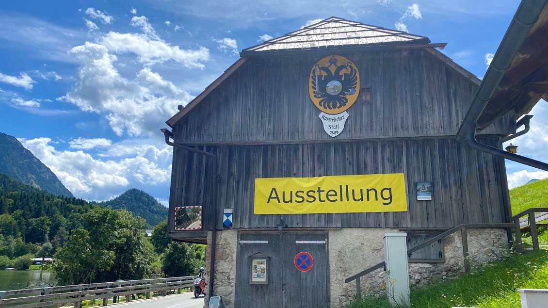 Ein Holzgebäude mit dem Schild "Ausstellung" und dem Wappen darüber. Im Hintergrund sind grüne Hügel und ein blauer Himmel mit Wolken zu sehen. | © TVB Ausseerland Salzkammergut/Bettina Scheck