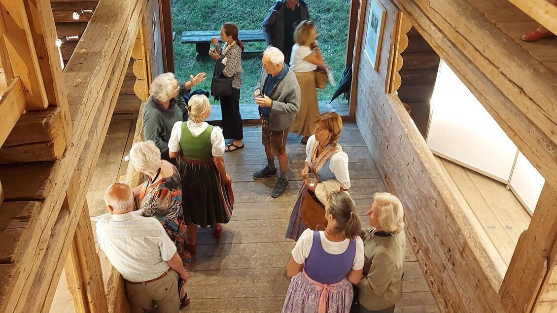 Eine Gruppe von Menschen in traditionellen Trachten steht in einem rustikalen Holzgebäude. Einige unterhalten sich lebhaft, während andere entspannt stehen und den Raum betrachten. | © Kulturelle ARGE Grundlsee