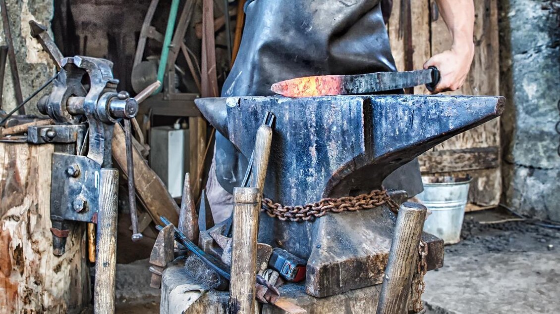 Ein Schmied arbeitet an einem Amboss und formt glühendes Metall. Die Werkstatt hat eine rustikale und handwerkliche Atmosphäre. | © Rudi Ferder