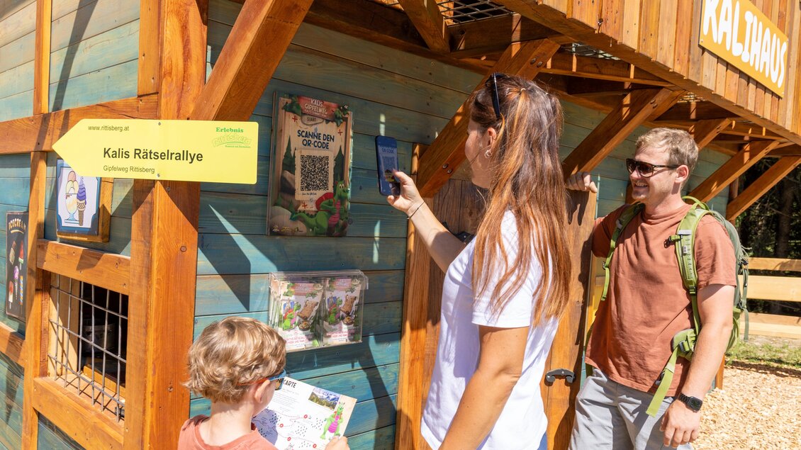 Eine Familie besucht eine interaktive Station im Freien. Ein Kind schaut auf eine Karte, während die Mutter Informationen scannt und der Vater daneben steht. | © Rittisberg