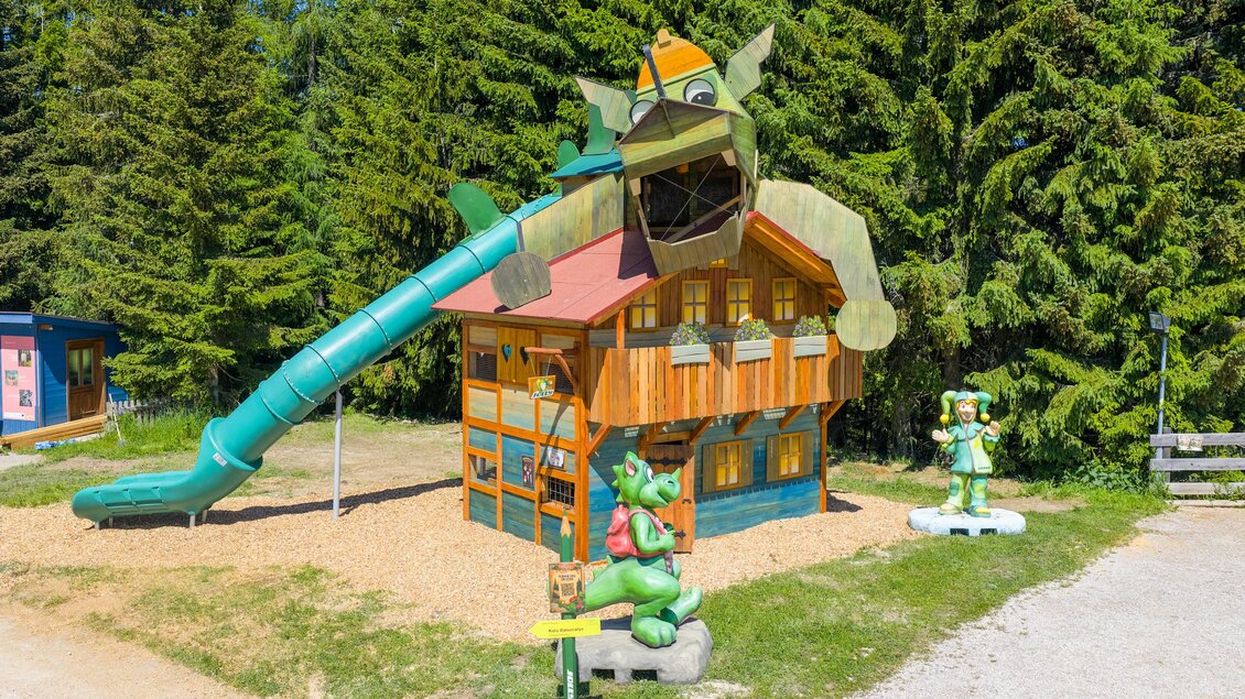 Ein bunter Spielplatz mit einem großen Spielhaus und einer Rutsche. Umgeben von hohen Bäumen bietet er viel Platz zum Spielen. | © Rittisberg