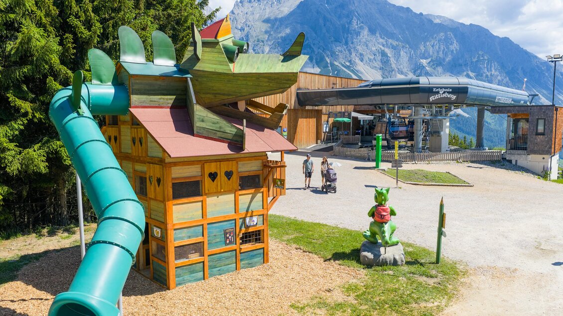 Ein bunter Spielplatz mit einer Drachenrutsche und einem Spielhaus befindet sich vor einer malerischen Berglandschaft. Im Hintergrund ist ein Gebäude sichtbar, das zu einem Freizeitbereich gehört. | © Rittisberg