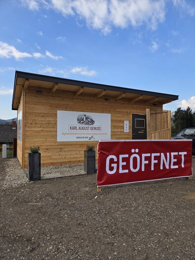 A wooden building with a sign that reads "Karl August Genuss". In front of the building hangs a large banner with the inscription "OPEN". | © Karl August