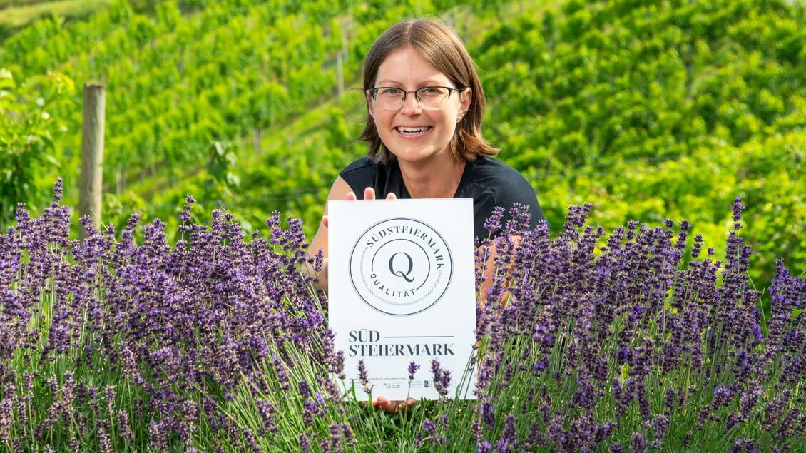 Eine lächelnde Frau steht zwischen lila Lavendelblüten und hält ein Schild mit der Aufschrift "Südsteiermark". Im Hintergrund sind grüne Weinberge zu sehen. | © RMSW GmbH-webquartier