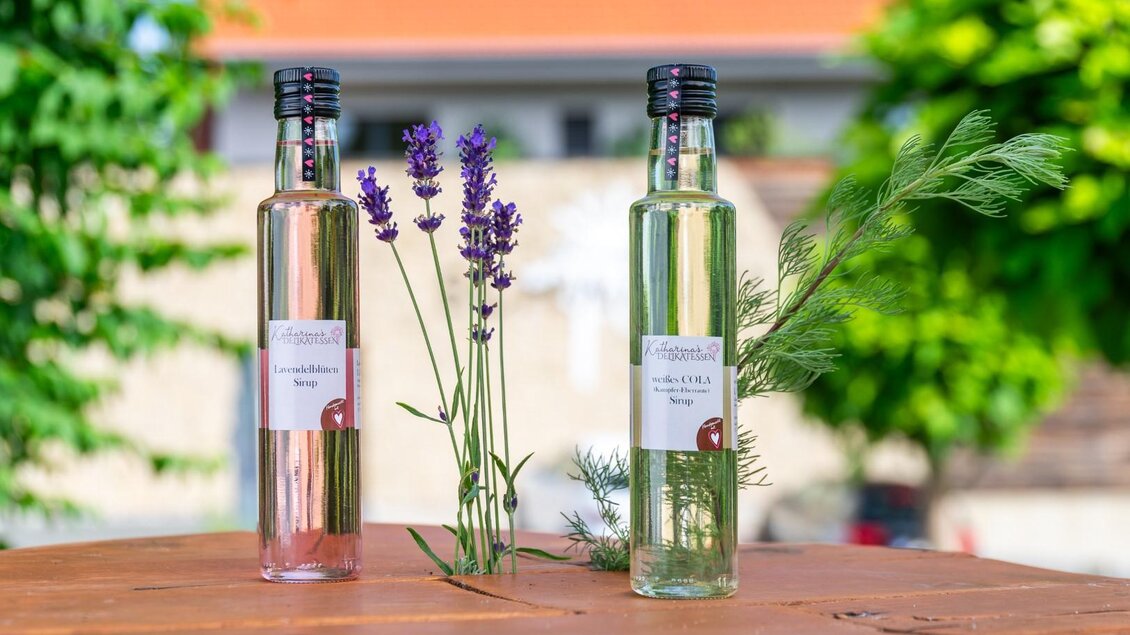 Zwei Flaschen aromatischer Likör stehen auf einem Holztisch. Daneben sind frische Kräuter und eine Lavendelblume arrangiert. | © RMSW GmbH-webquartier