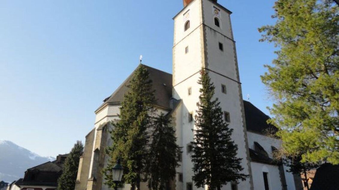 Eine historische Kirche mit einem hohen Turm und umgebenden Bäumen. Im Hintergrund sind die Berge zu sehen.