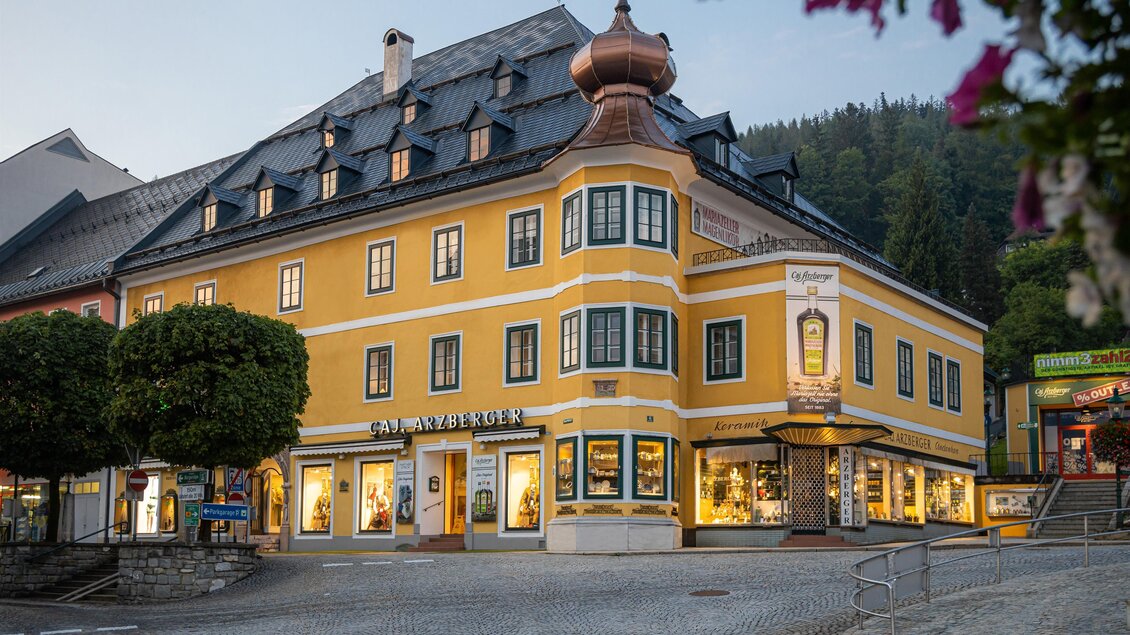 Ein buntes, gelbes Gebäude mit einem ausgefallenen Dach und traditionellen Fenstern. Es befindet sich in einer malerischen Umgebung mit grünen Bäumen und Bergen im Hintergrund. | © Arzberger Likörmanufaktur