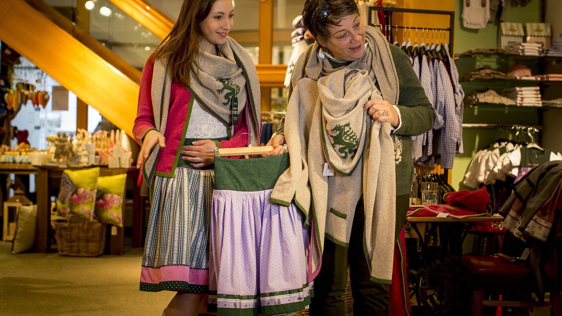 Zwei Frauen stehen in einem Geschäft und präsentieren traditionelle Kleidung. Die eine hält einen Rock und beide tragen Schals im typischen Stil. | © TV Hochsteiermark, Tom Lamm
