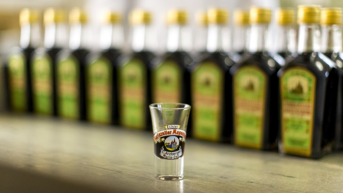 Ein Schussglas steht im Vordergrund, während im Hintergrund mehrere Flaschen mit einem dunklen Getränk sichtbar sind. Die Flaschen haben gelbe Kappen und Etiketten mit grünen Details. | © TV Hochsteiermark, Tom Lamm