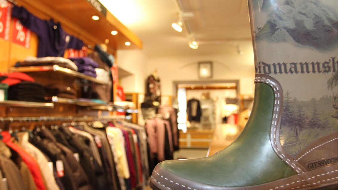 Ein Gummistiefel steht im Vordergrund, während der Hintergrund mit Kleidung in einem Geschäft gefüllt ist. Es ist eine gemütliche Boutique-Atmosphäre zu sehen.