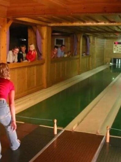 Eine Bowlingbahn in einem Holzraum mit einer Spielerin, die einen Ball wirft. Im Hintergrund beobachten mehrere Personen das Spiel. | © Reisslerhof
