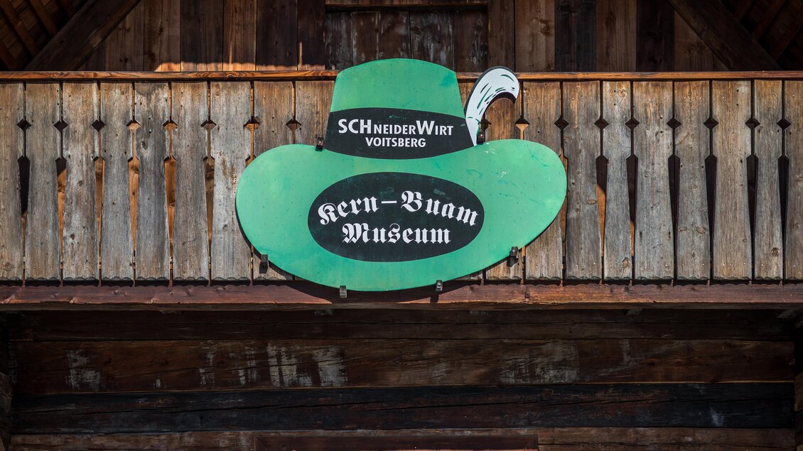Grünes Schild des Kern Buam Museums in Voitsberg am Holzbalkon des Schneiderwirts mit der Aufschrift „Kern-Buam Museum“  | © DieAbbilderei