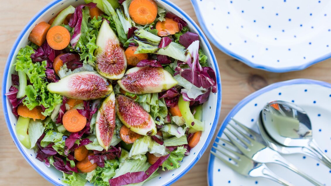 Ein bunter Salat mit frischem Gemüse und Feigen in einer Schüssel. Neben der Schüssel stehen zwei Teller und Besteck. | © Michaela Lorber