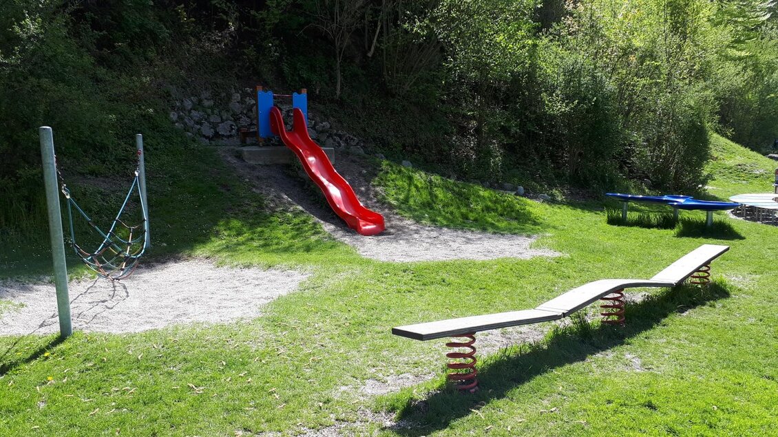 <p>Der Spielplatz im Kurpark in Bad Aussee mit einer roten Rutsche, einer Wippe und Schaukel. Der Bereich ist von grünem Gras und Bäumen umgeben.</p> | © Stadtgemeinde Bad Aussee/Ziermayer