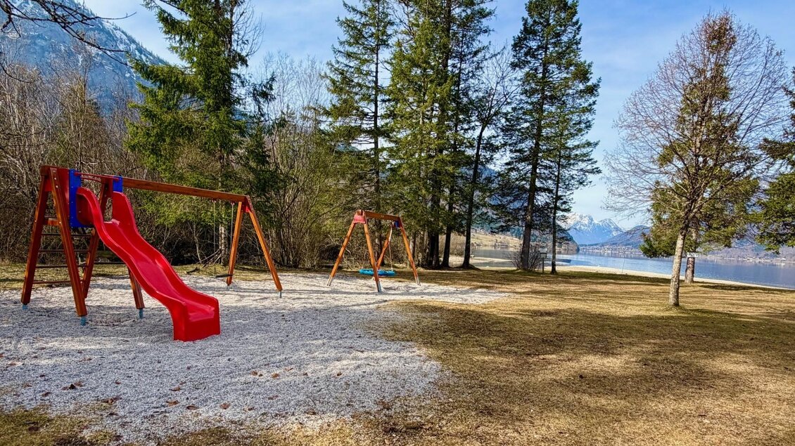 Ein Spielplatz mit einer roten Rutsche und Schaukeln steht im Grünen. Im Hintergrund sind Bäume und ein ruhiger See zu sehen. | © TVB Ausseerland