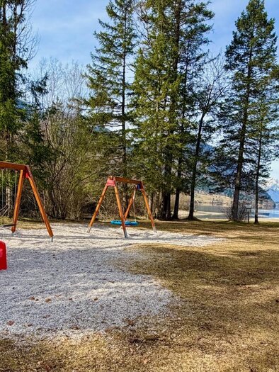 Ein Spielplatz mit einer roten Rutsche und Schaukeln steht im Grünen. Im Hintergrund sind Bäume und ein ruhiger See zu sehen. | © TVB Ausseerland