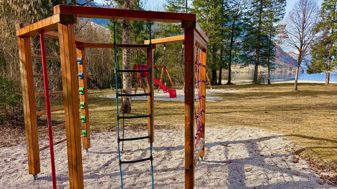 Ein Spielplatz mit Klettergerüst und Rutsche steht in einer ruhigen Parklandschaft. Im Hintergrund sind Bäume und ein See sichtbar. | © TVB Ausseerland