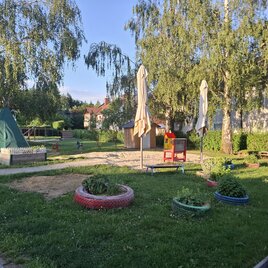 Ein Spielplatz mit Sandfläche, einem Spielhaus und verschiedenen Sitzgelegenheiten. Der Garten ist von Bäumen umgeben und hat bunte Blumentöpfe. | © Gerlinde Rath