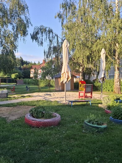 Ein Spielplatz mit Sandfläche, einem Spielhaus und verschiedenen Sitzgelegenheiten. Der Garten ist von Bäumen umgeben und hat bunte Blumentöpfe. | © Gerlinde Rath
