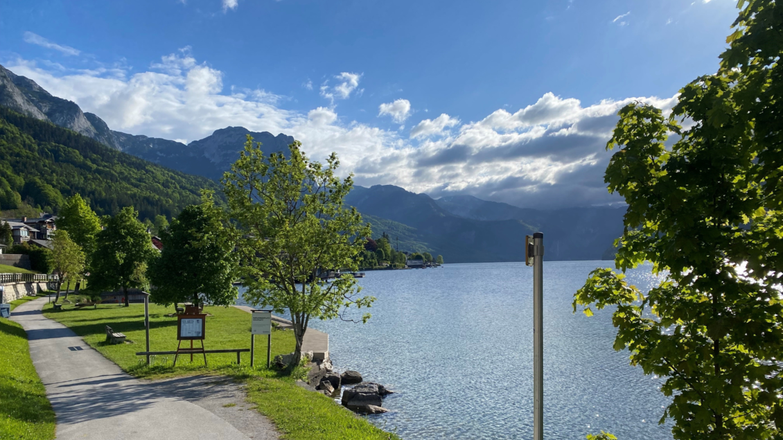<p>Der schöne Grundlsee umgeben von Bergen und Bäumen. Unten am Ufer kann der Spielplatz gefunden werden. Der Himmel ist teilweise bewölkt und das Wasser glitzert in der Sonne.</p> | © Bettina Scheck