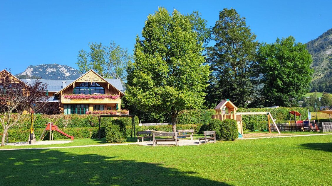 Ein schöner Park mit viel grünem Gras und großen Bäumen. Im Hintergrund steht ein Spielplatz mit Schaukel und Rutsche. | © Petra Kirchschlager