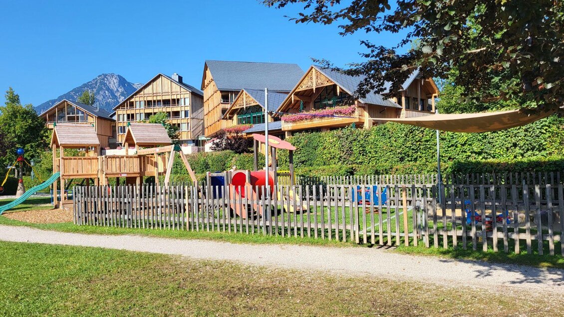 Ein Spielplatz mit Holzspielgeräten und einer feinen Rasenfläche. Im Hintergrund sind moderne Häuser und ein beeindruckender Berg zu sehen. | © Petra Kirchschlager