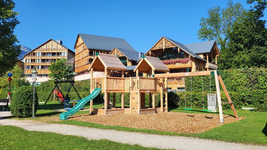 Ein schöner Spielplatz mit einem Spielhaus, einer Rutsche und Schaukeln. Im Hintergrund stehen moderne Gebäude und viel Grün. | © Petra Kirchschlager