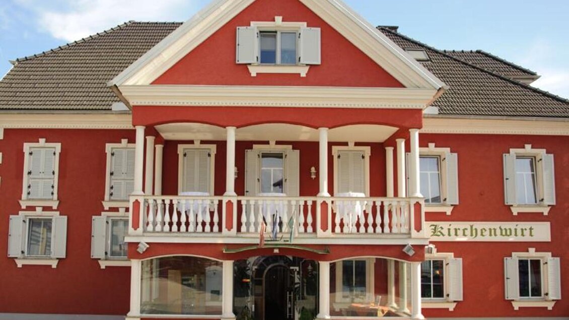 Ein rotes Gebäude mit weißem Balkon und Fenstern. Es steht der Schriftzug "Kirchenwirt" an der Fassade. | © Gasthaus Draxler