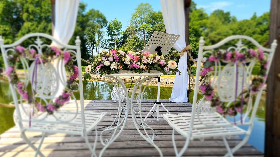 Ein romantischer Pavillon am Wasser mit einem weißen Tisch und zwei Stühlen. Der Tisch ist mit Blumen geschmückt und die Umgebung ist grün und einladend. | © Gasthaus Draxler