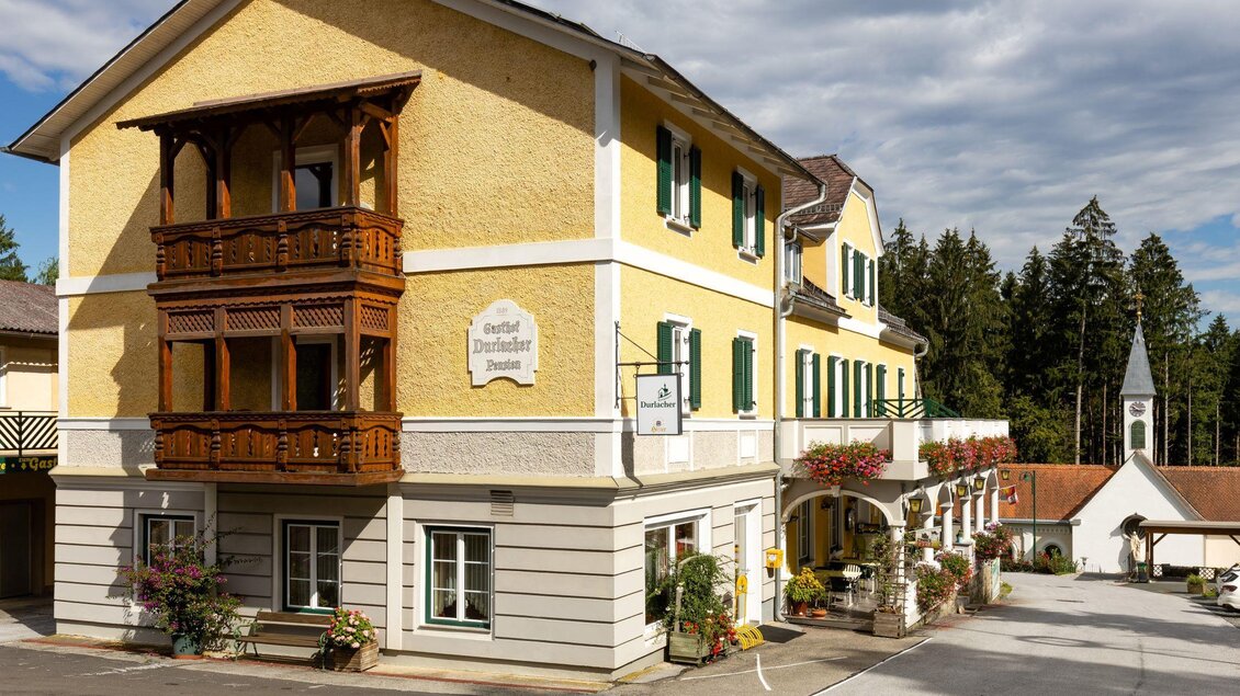 Ein charmantes gelbes Gebäude mit Holzbalustrade und grünen Fenstern. Im Hintergrund ist ein Kirchturm und Bäume zu sehen. | © Gasthof Durlacher