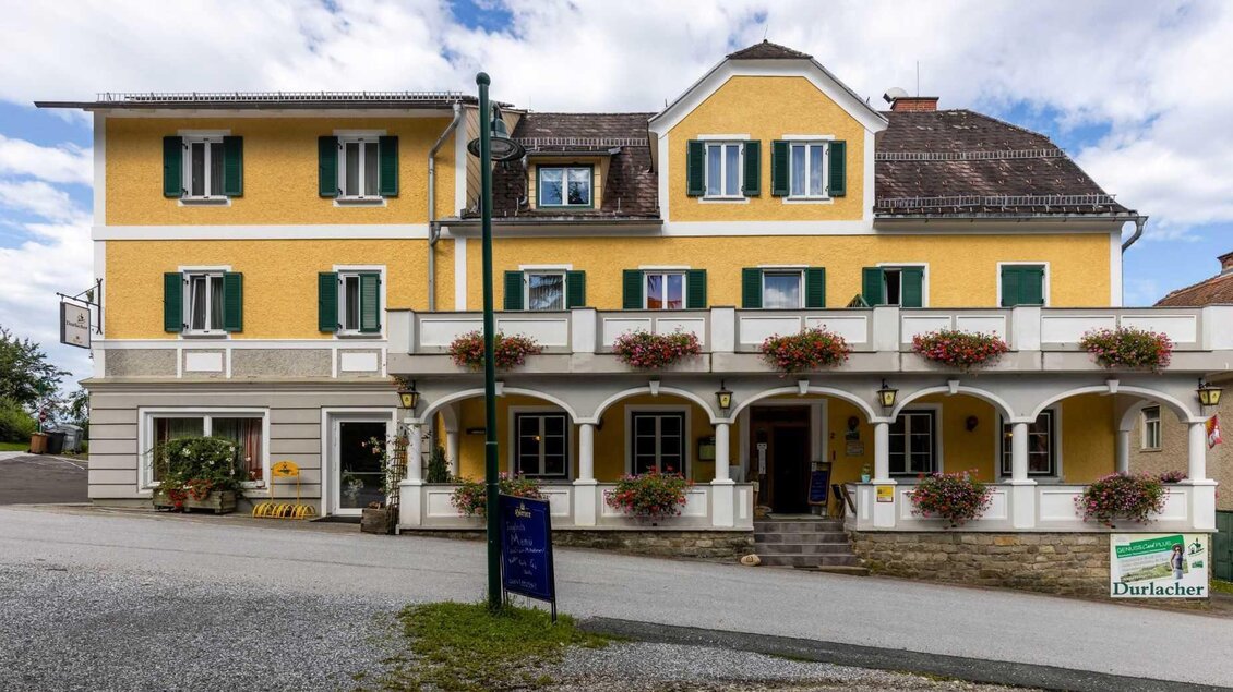 Ein gelbes Gebäude mit grünen Fensterläden und Blumen an den Fenstern. Vor dem Haus befinden sich ein paar Parkplätze und eine klare Himmelsszenerie. | © Gasthof Durlacher