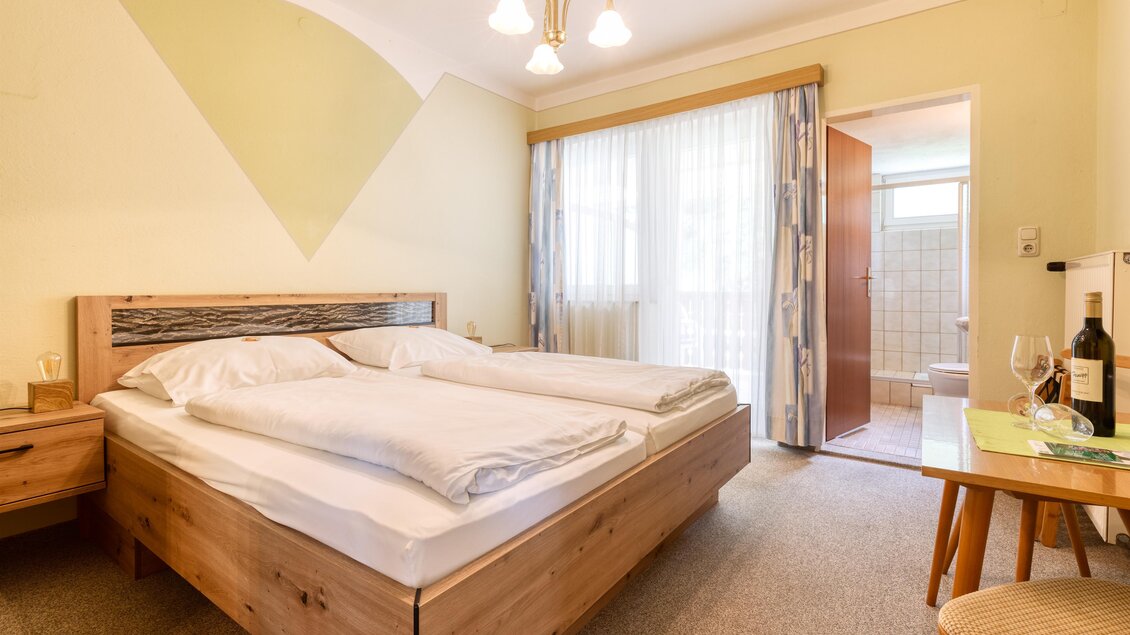 Ein gemütliches Schlafzimmer mit einem Doppelbett und Holzdetails. Es gibt große Fenster, die viel Tageslicht hereinlassen, sowie einen Zugang zu einem Badezimmer. | © Gasthof Durlacher