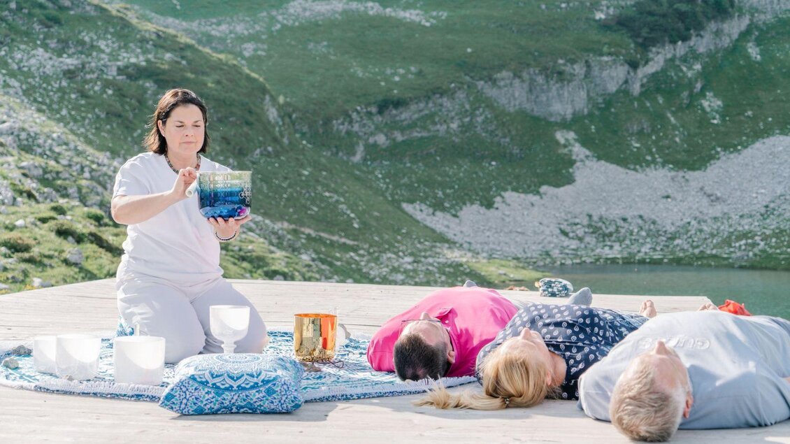 <p>Stephanie Bor leitet eine Klangschalensitzung in den Bergen des Ausseerlandes. Mehrere Personen liegen auf einer Plattform und genießen die friedliche Landschaft.</p> | © Lebensbilder, www.deine-lebensbilder.at/impressum/