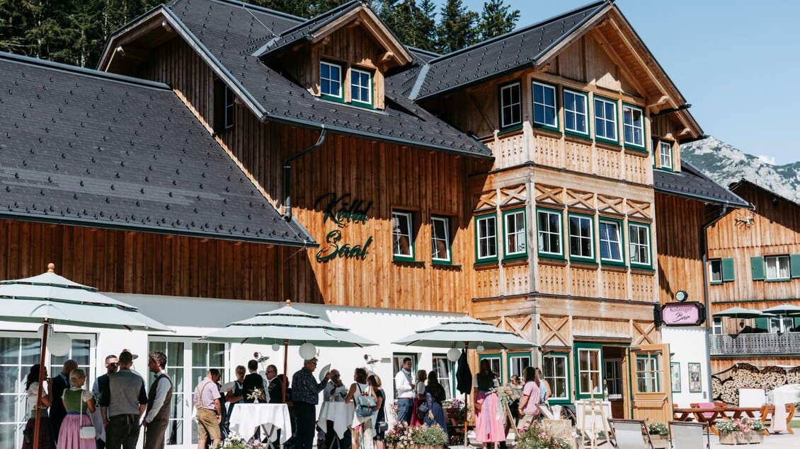 Ein traditionelles Holzgebäude mit mehreren Fenstern und einem einladenden Eingang. Im Vordergrund versammeln sich Gruppen von Menschen, die eine Veranstaltung im Freien genießen.