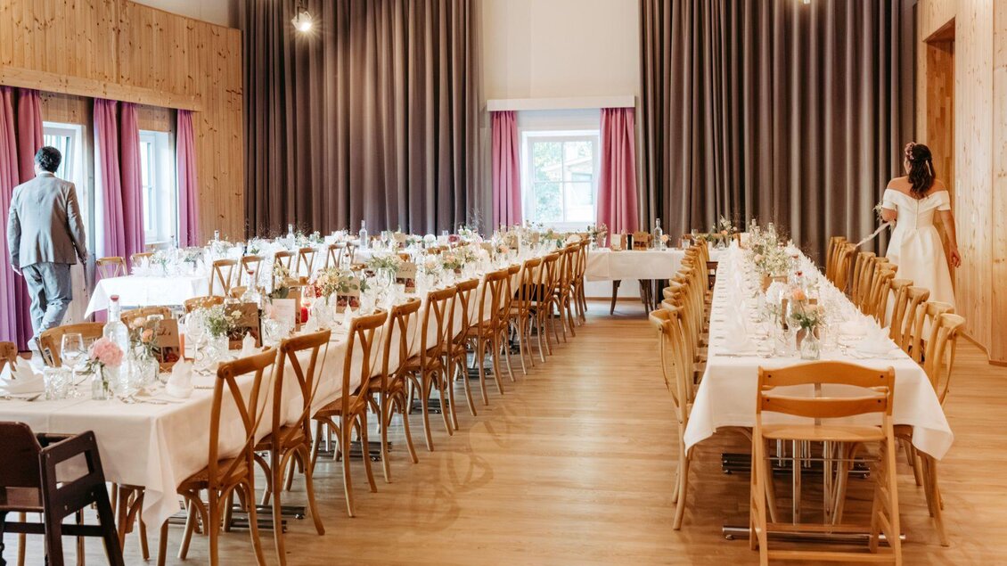 Ein festlich dekorierter Saal mit langen Tischen und eleganten Tischgedecken. Eine Hochzeitsfeier wird vorbereitet, mit einer Braut und Gästen im Hintergrund. | © Narzissendorf Zloam www.zloam.at