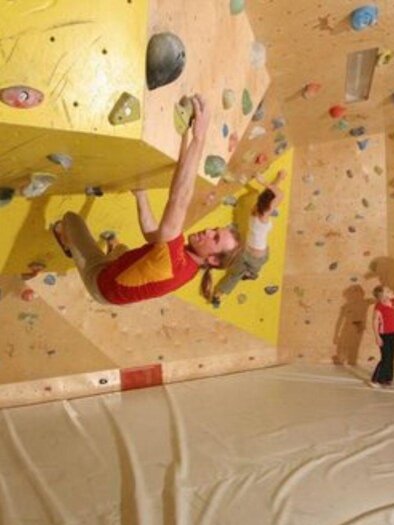 Eine Kletterwand mit bunten Griffen in einer Kletterhalle. Ein Kletterer hängt an der Wand, während zwei Zuschauer zuschauen. | © Mirja Geh
