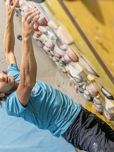 Ein Kletterer hängt an einer Boulderwand und konzentriert sich auf seinen nächsten Griff. Die Wand ist mit bunten Griffen ausgestattet, und der Hintergrund ist in blassem Blau. | © Thomas Sattler