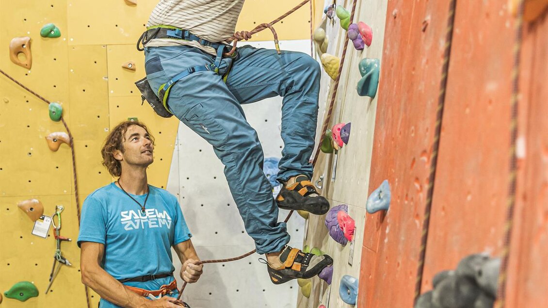 Ein Kletterer steigt an einer Indoor-Kletterwand, während sein Partner ihn sichert. Die Wände sind bunt gestaltet und mit verschiedenen Klettergriffen ausgestattet. | © Thomas Sattler